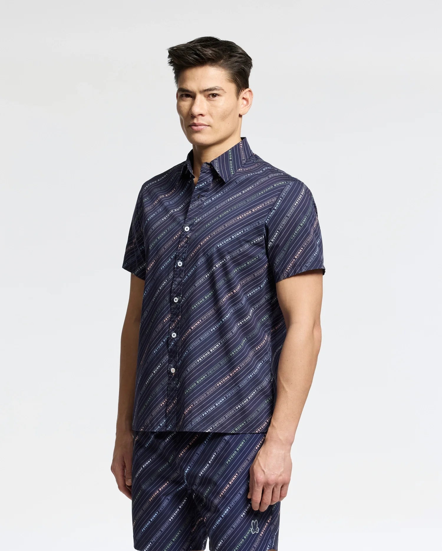 A person stands against a white background in a Psycho Bunny MENS RENMARK STRETCH POPLIN WOVEN SHIRT (B6Q629F200), featuring navy blue stretch poplin with diagonal white stripes. With short, dark hair, they pair the shirt with matching pants.