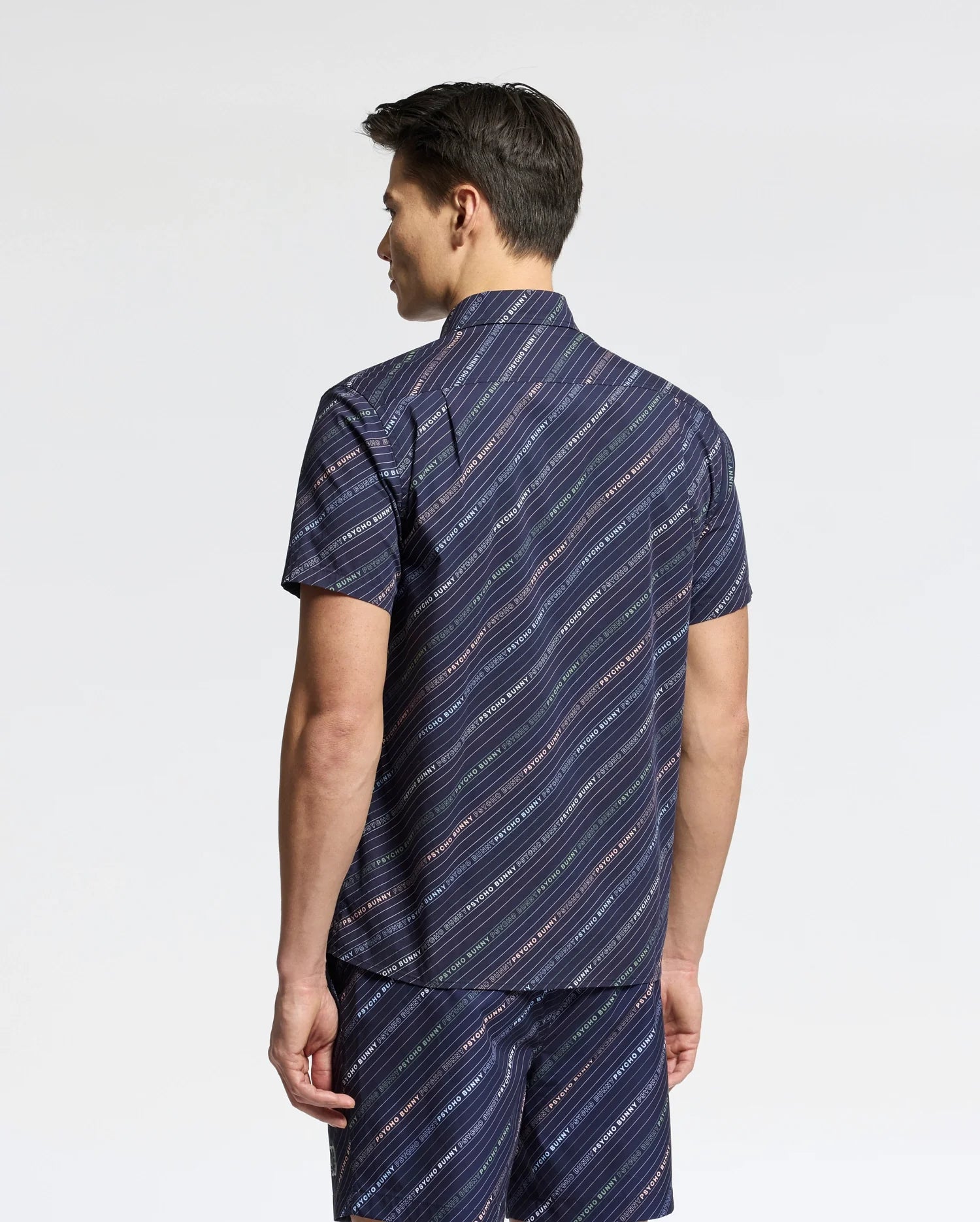 A man is seen from behind in a Psycho Bunny MENS RENMARK STRETCH POPLIN WOVEN SHIRT (B6Q629F200) and matching shorts, featuring a blue logo-striped pattern. The background is plain white.