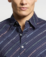 A person sports the MENS RENMARK STRETCH POPLIN WOVEN SHIRT by Psycho Bunny, showcasing a colorful logo-striped pattern. Crafted from stretch poplin for comfort, it features white buttons and a partially visible face among the vibrant stripes.
