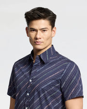 A person stands against a stark white background, wearing Psycho Bunny's MENS RENMARK STRETCH POPLIN WOVEN SHIRT - B6Q629F200. This dark blue button-up features a logo-striped pattern and text detail, effortlessly blending style with comfort, perfect for any occasion.