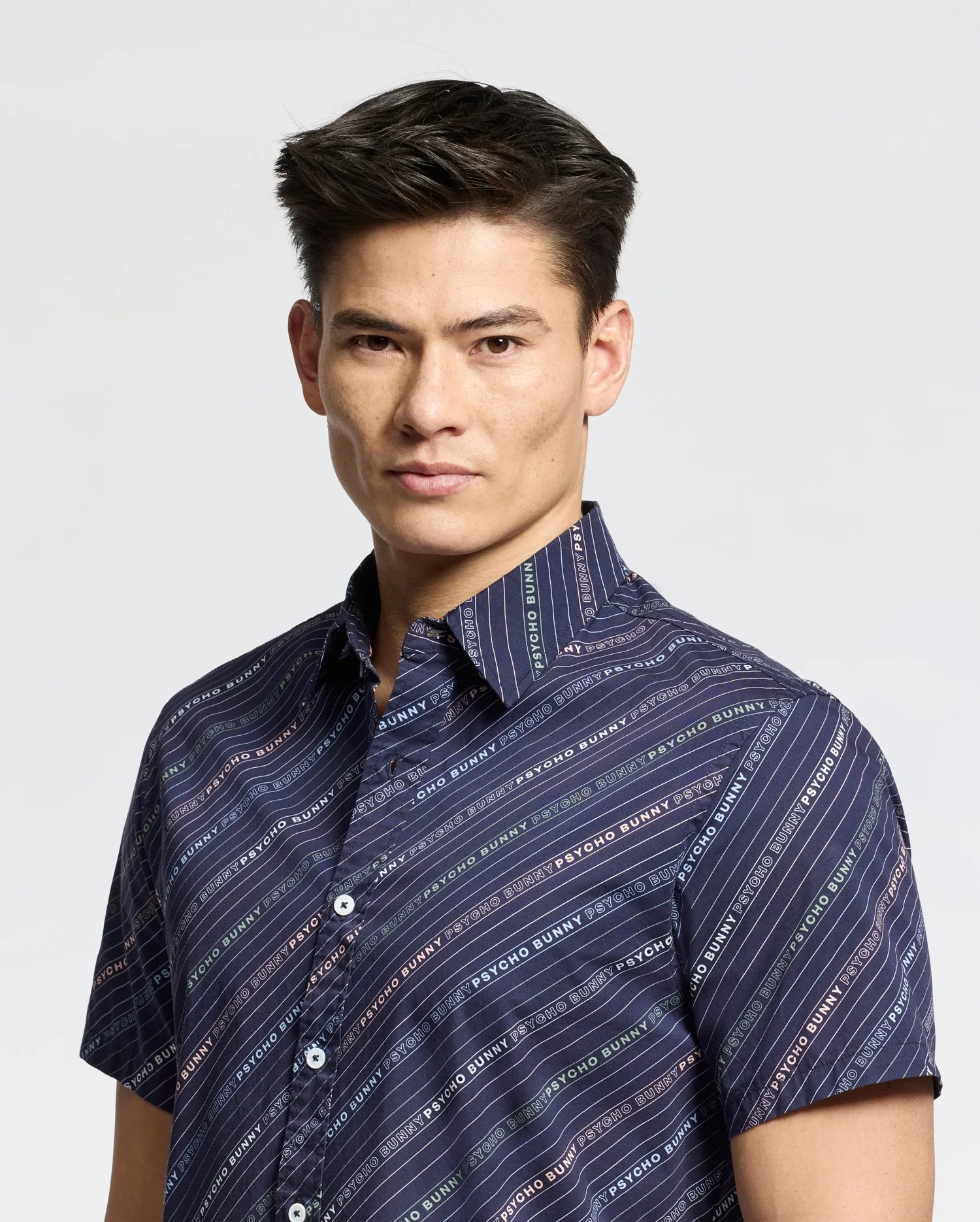 A person stands against a stark white background, wearing Psycho Bunny's MENS RENMARK STRETCH POPLIN WOVEN SHIRT - B6Q629F200. This dark blue button-up features a logo-striped pattern and text detail, effortlessly blending style with comfort, perfect for any occasion.