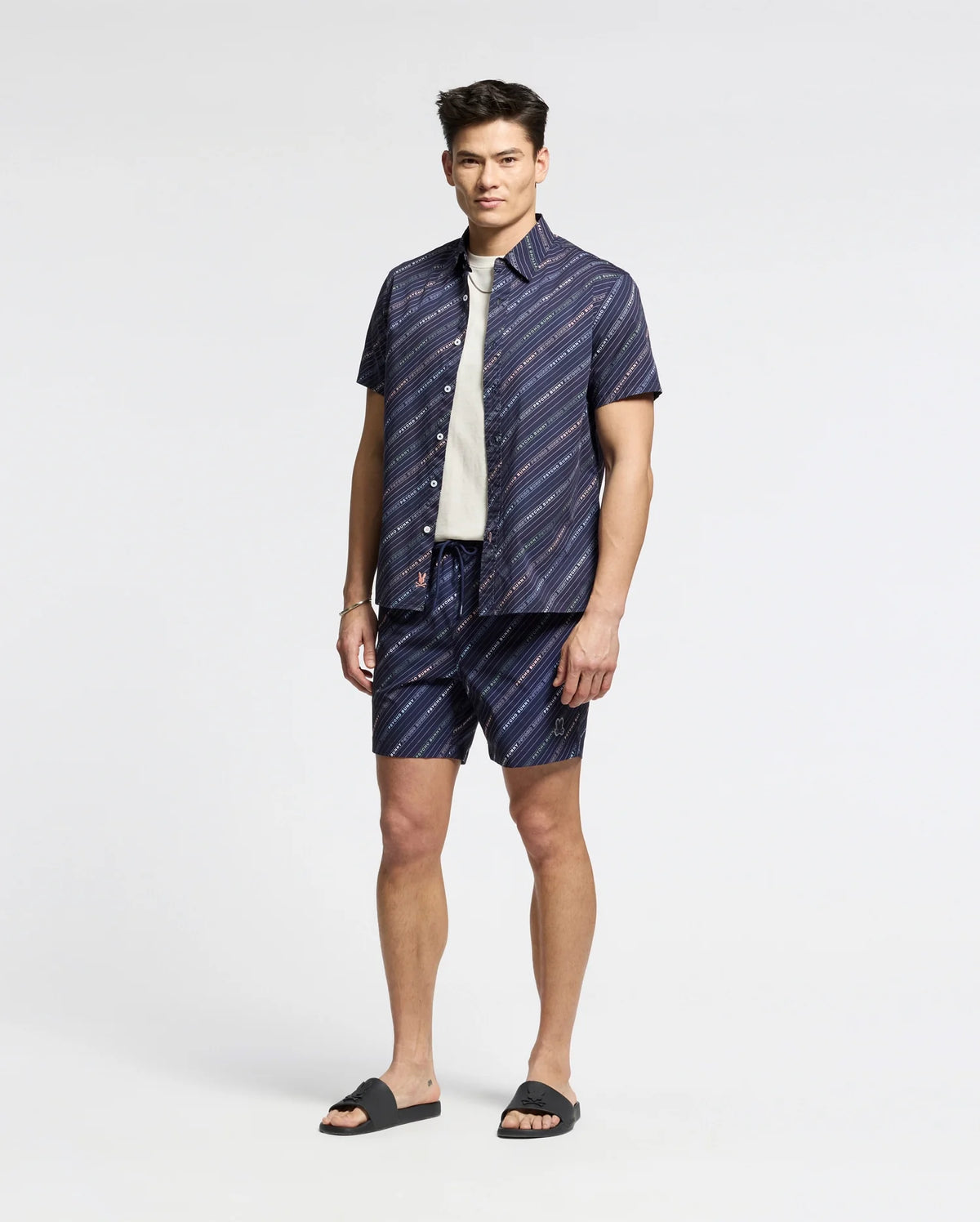 Someone stands against a plain background, wearing a navy Psycho Bunny short-sleeve button-up shirt and matching logo-striped shorts. A white tee peeks out underneath, paired with black slides for a relaxed yet polished look. MENS RENMARK STRETCH POPLIN WOVEN SHIRT - B6Q629F200.