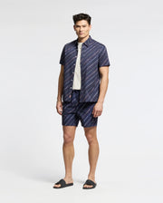 Someone stands against a plain background, wearing a navy Psycho Bunny short-sleeve button-up shirt and matching logo-striped shorts. A white tee peeks out underneath, paired with black slides for a relaxed yet polished look. MENS RENMARK STRETCH POPLIN WOVEN SHIRT - B6Q629F200.