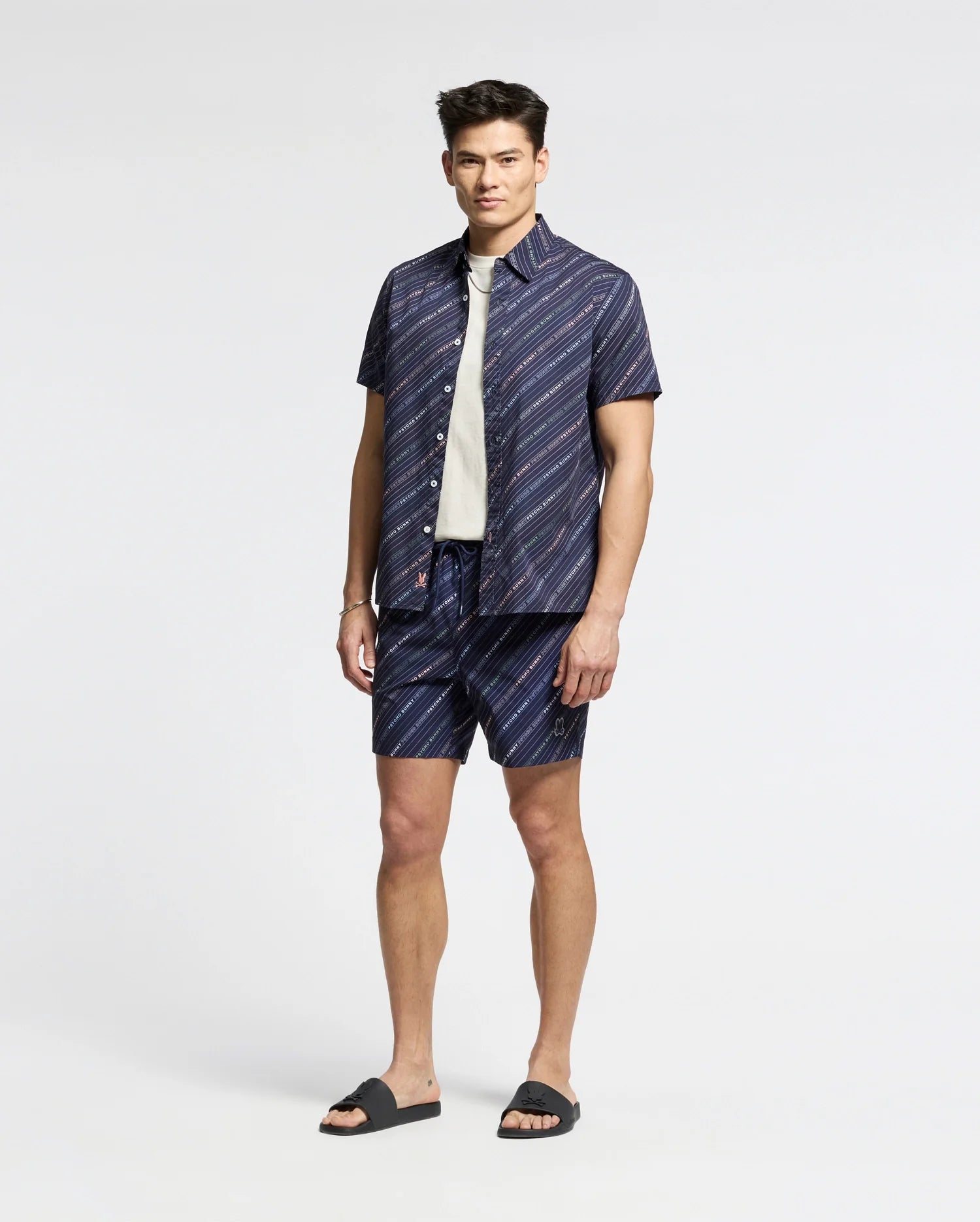 Someone stands against a plain background, wearing a navy Psycho Bunny short-sleeve button-up shirt and matching logo-striped shorts. A white tee peeks out underneath, paired with black slides for a relaxed yet polished look. MENS RENMARK STRETCH POPLIN WOVEN SHIRT - B6Q629F200.