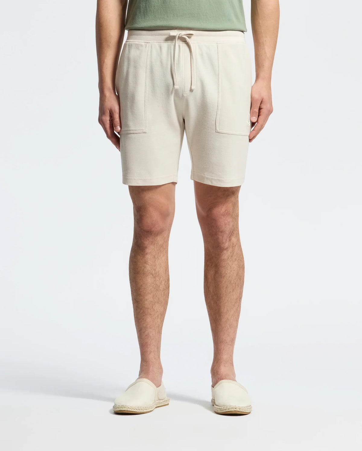 Wearing Psycho Bunny's MENS TAYLOR LIGHT PIQUE SWEATSHORT in beige with drawstrings and front pockets, paired with a green shirt, and light beige slip-on shoes made from lightweight Pima cotton, the person strikes a casual-chic pose against a plain white background.