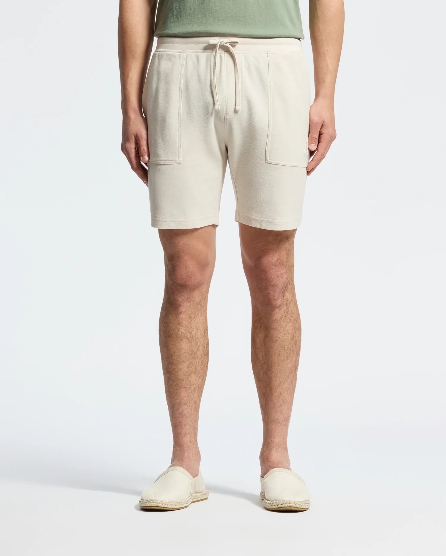 Wearing Psycho Bunny's MENS TAYLOR LIGHT PIQUE SWEATSHORT in beige with drawstrings and front pockets, paired with a green shirt, and light beige slip-on shoes made from lightweight Pima cotton, the person strikes a casual-chic pose against a plain white background.
