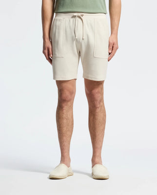 Wearing Psycho Bunny's MENS TAYLOR LIGHT PIQUE SWEATSHORT in beige with drawstrings and front pockets, paired with a green shirt, and light beige slip-on shoes made from lightweight Pima cotton, the person strikes a casual-chic pose against a plain white background.