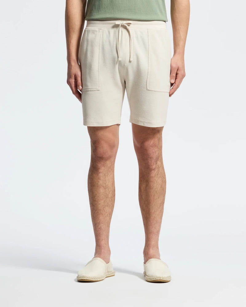 Wearing Psycho Bunny's MENS TAYLOR LIGHT PIQUE SWEATSHORT in beige with drawstrings and front pockets, paired with a green shirt, and light beige slip-on shoes made from lightweight Pima cotton, the person strikes a casual-chic pose against a plain white background.
