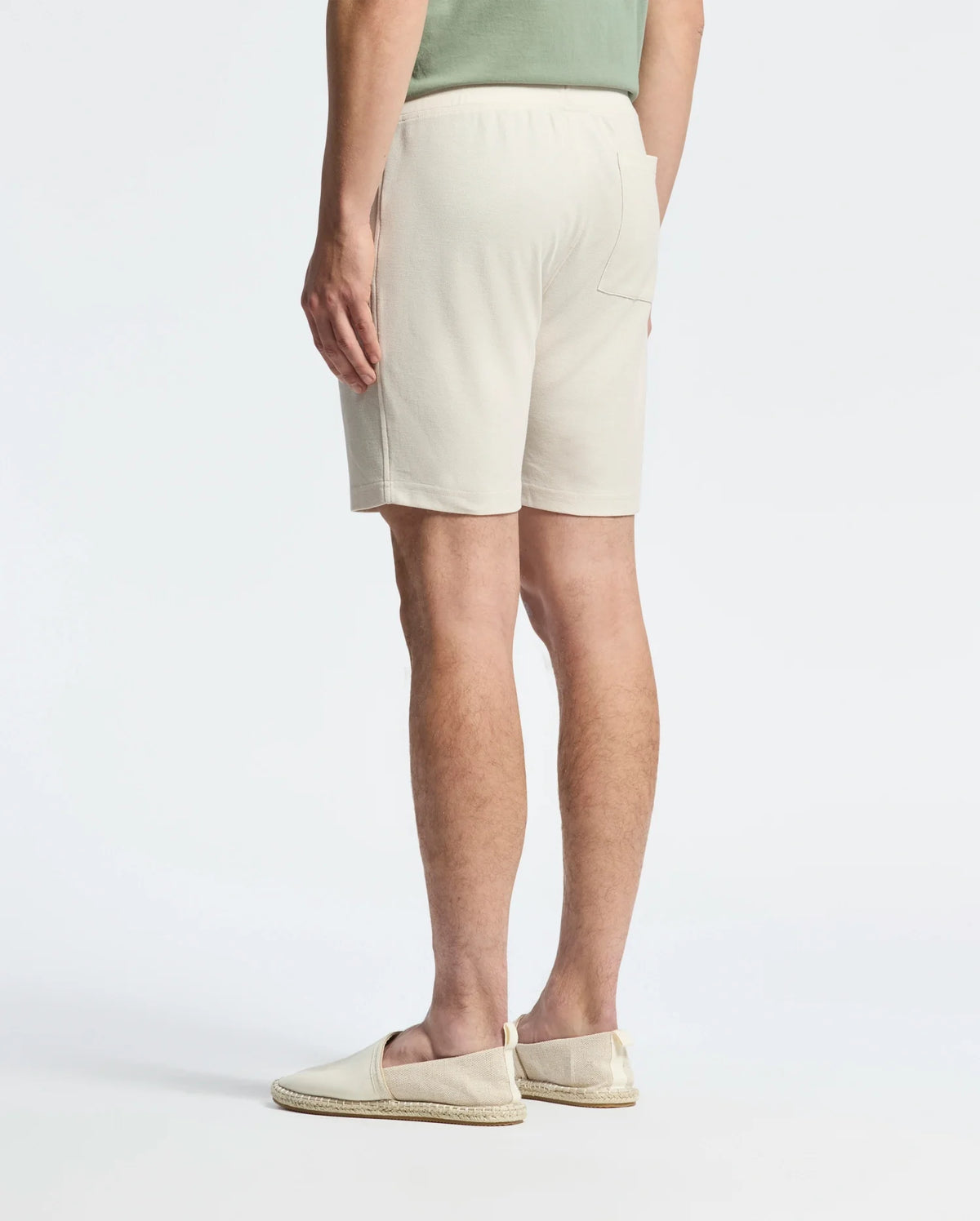 A person in a casual-chic outfit featuring a light green shirt and Psycho Bunny's Taylor Light Pique Sweatshort in beige, crafted from lightweight Pima cotton, stands with their back to the camera. They finish the look with beige slip-on shoes against a plain white backdrop.