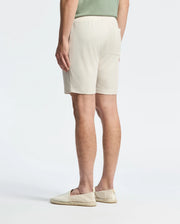 A person in a casual-chic outfit featuring a light green shirt and Psycho Bunny's Taylor Light Pique Sweatshort in beige, crafted from lightweight Pima cotton, stands with their back to the camera. They finish the look with beige slip-on shoes against a plain white backdrop.