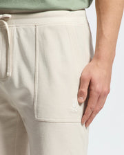 A close-up shows someone in cream MENS TAYLOR LIGHT PIQUE SWEATSHORT by Psycho Bunny, crafted from lightweight Pima cotton with visible pocket stitching and an embroidered rabbit logo, resting one hand on the shorts while wearing a light green shirt.