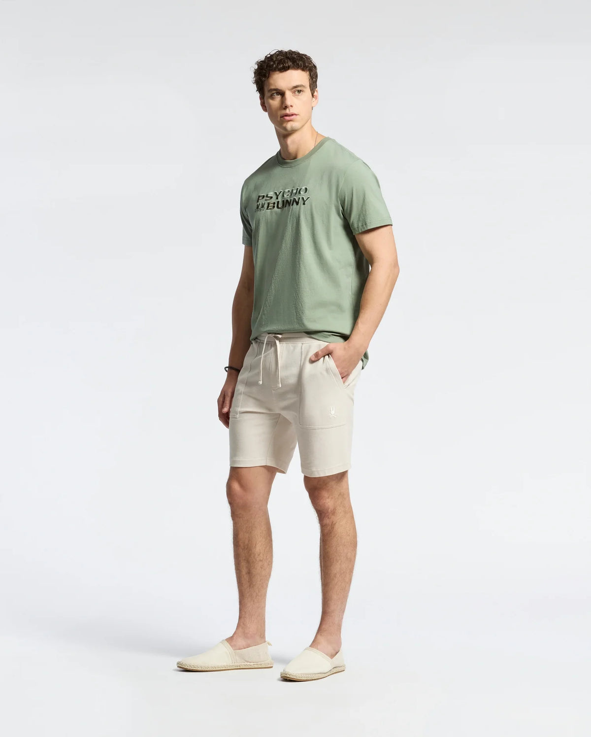A man wearing a light green, lightweight Pima cotton t-shirt, Psycho Bunny MENS TAYLOR LIGHT PIQUE SWEATSHORTS in cream, and slip-on shoes stands against a plain background with his hands in his pockets, exuding casual-chic ease.