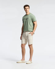 A man wearing a light green, lightweight Pima cotton t-shirt, Psycho Bunny MENS TAYLOR LIGHT PIQUE SWEATSHORTS in cream, and slip-on shoes stands against a plain background with his hands in his pockets, exuding casual-chic ease.