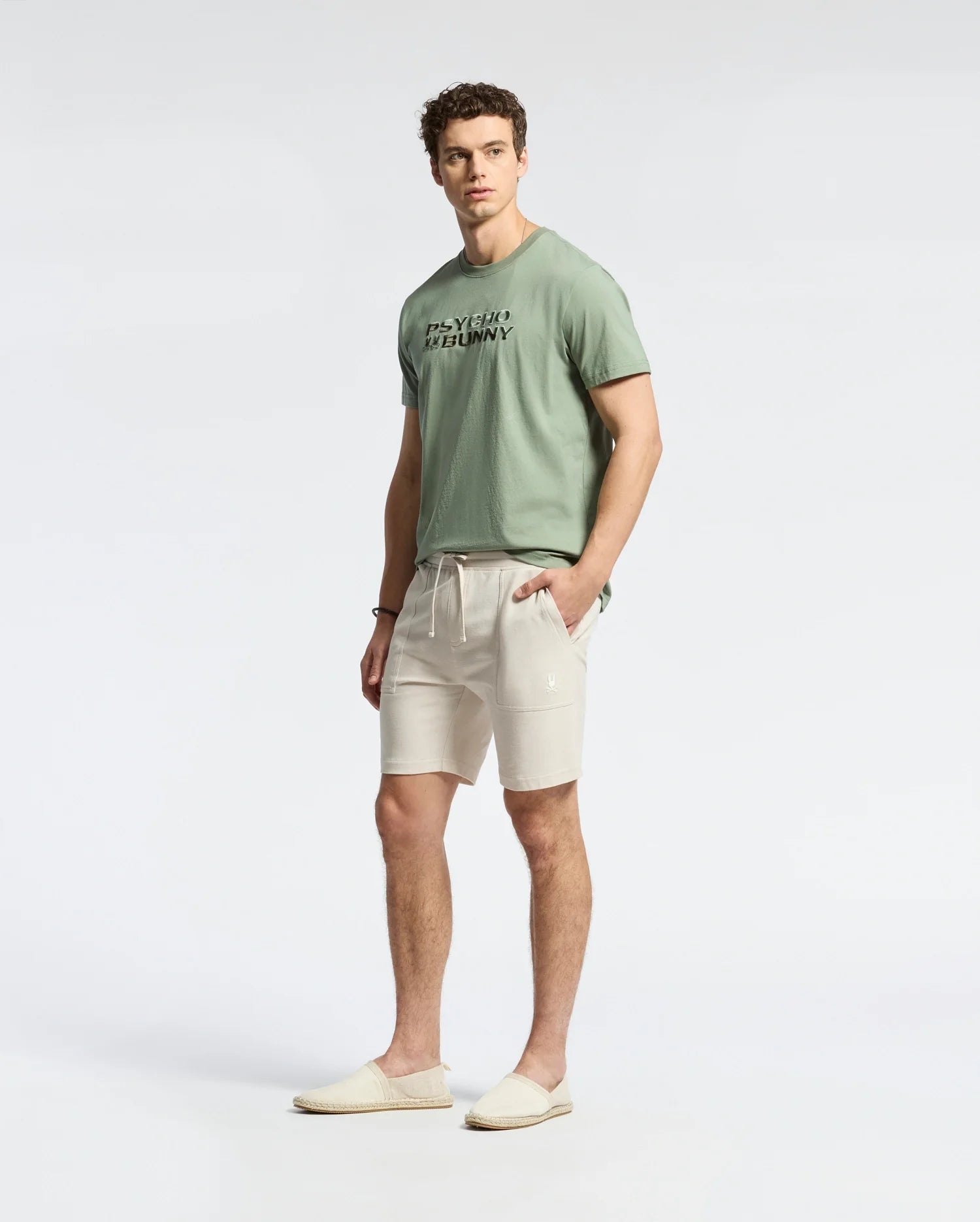 A man wearing a light green, lightweight Pima cotton t-shirt, Psycho Bunny MENS TAYLOR LIGHT PIQUE SWEATSHORTS in cream, and slip-on shoes stands against a plain background with his hands in his pockets, exuding casual-chic ease.