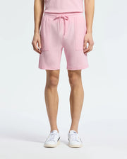 A person wears the chic Psycho Bunny MENS TAYLOR LIGHT PIQUE SWEATSHORT in pink, made of lightweight Pima cotton with drawstrings and large pockets. They complete the look with white sneakers featuring a blue logo against a plain white background.