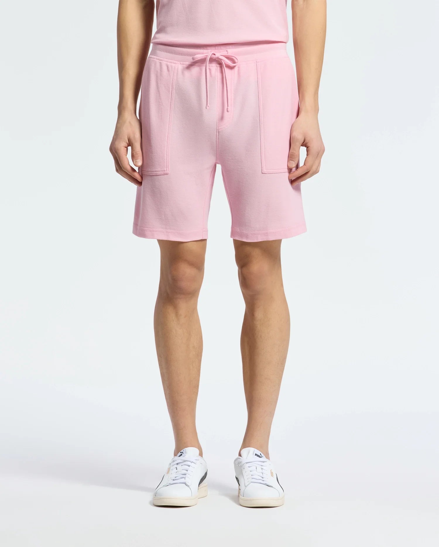 A person wears the chic Psycho Bunny MENS TAYLOR LIGHT PIQUE SWEATSHORT in pink, made of lightweight Pima cotton with drawstrings and large pockets. They complete the look with white sneakers featuring a blue logo against a plain white background.