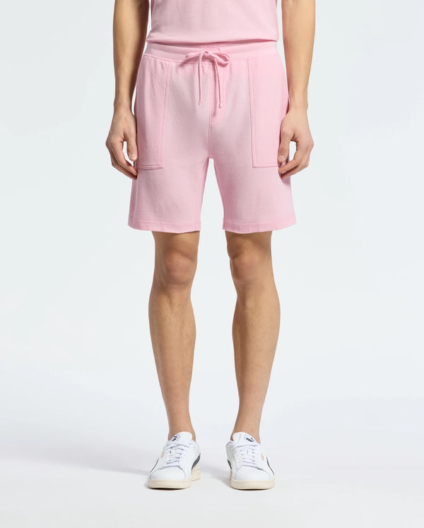 A person wears the chic Psycho Bunny MENS TAYLOR LIGHT PIQUE SWEATSHORT in pink, made of lightweight Pima cotton with drawstrings and large pockets. They complete the look with white sneakers featuring a blue logo against a plain white background.