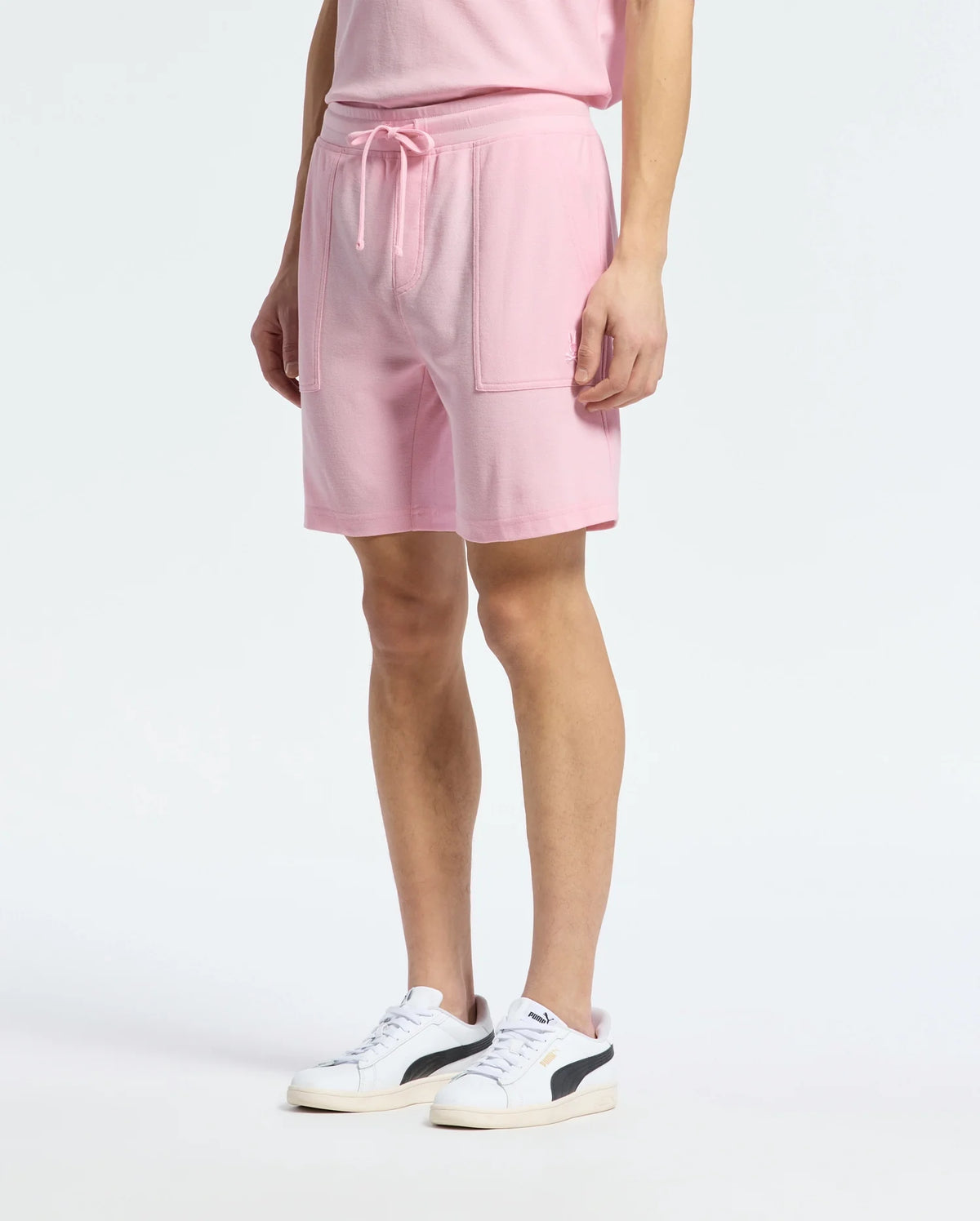 A person effortlessly embodies casual-chic style wearing the Psycho Bunny MENS TAYLOR LIGHT PIQUE SWEATSHORT in light pink, teamed with a matching lightweight t-shirt and white sneakers accented in black.
