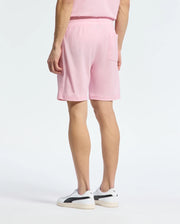 A person in a casual-chic pink Pima cotton outfit, featuring the Psycho Bunny MENS TAYLOR LIGHT PIQUE SWEATSHORT with a back pocket, stands with their back to the camera. They wear white sneakers with black accents against a plain white background.