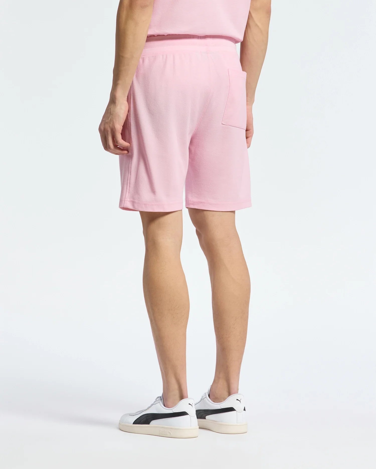 A person in a casual-chic pink Pima cotton outfit, featuring the Psycho Bunny MENS TAYLOR LIGHT PIQUE SWEATSHORT with a back pocket, stands with their back to the camera. They wear white sneakers with black accents against a plain white background.
