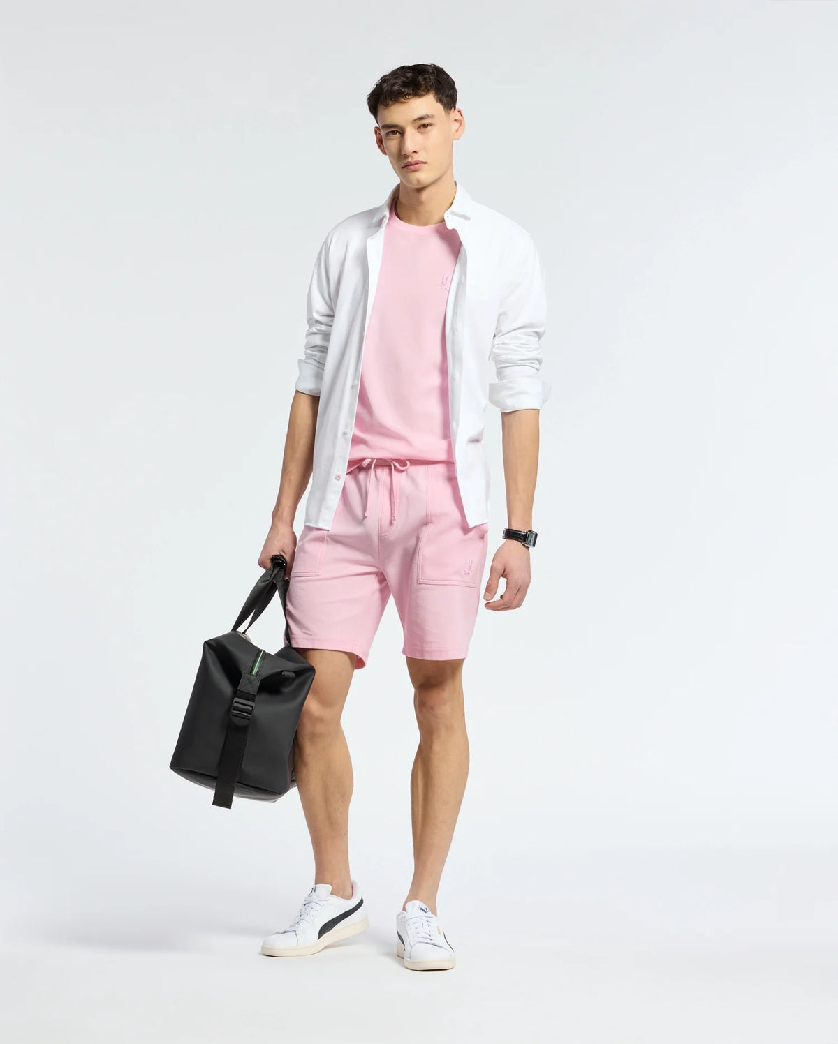 A person stands against a plain background, exuding casual-chic style in a white open shirt over a pink T-shirt and matching Psycho Bunny MENS TAYLOR LIGHT PIQUE SWEATSHORT. They hold a black bag in one hand and wear lightweight white sneakers.