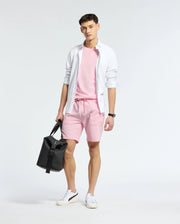 A person stands against a plain background, exuding casual-chic style in a white open shirt over a pink T-shirt and matching Psycho Bunny MENS TAYLOR LIGHT PIQUE SWEATSHORT. They hold a black bag in one hand and wear lightweight white sneakers.