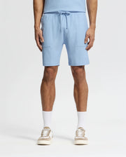 The individual sports Psycho Bunny's light blue Pima cotton MENS TAYLOR LIGHT PIQUE SWEATSHORT - B6R344F200 featuring a drawstring and patch pockets, pairing them with white socks and beige sneakers. This casual-chic style pops against the plain white background.