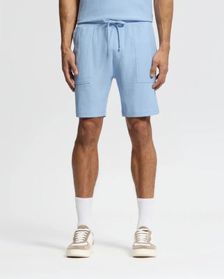 The individual sports Psycho Bunny's light blue Pima cotton MENS TAYLOR LIGHT PIQUE SWEATSHORT - B6R344F200 featuring a drawstring and patch pockets, pairing them with white socks and beige sneakers. This casual-chic style pops against the plain white background.