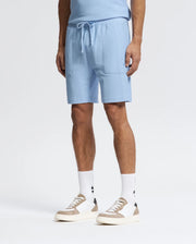 Wearing Psycho Bunny's MENS TAYLOR LIGHT PIQUE SWEATSHORTS and a matching blue shirt, paired with white socks featuring a black logo and white sneakers with beige and gray highlights, creates an ideal casual-chic look against the plain white backdrop.