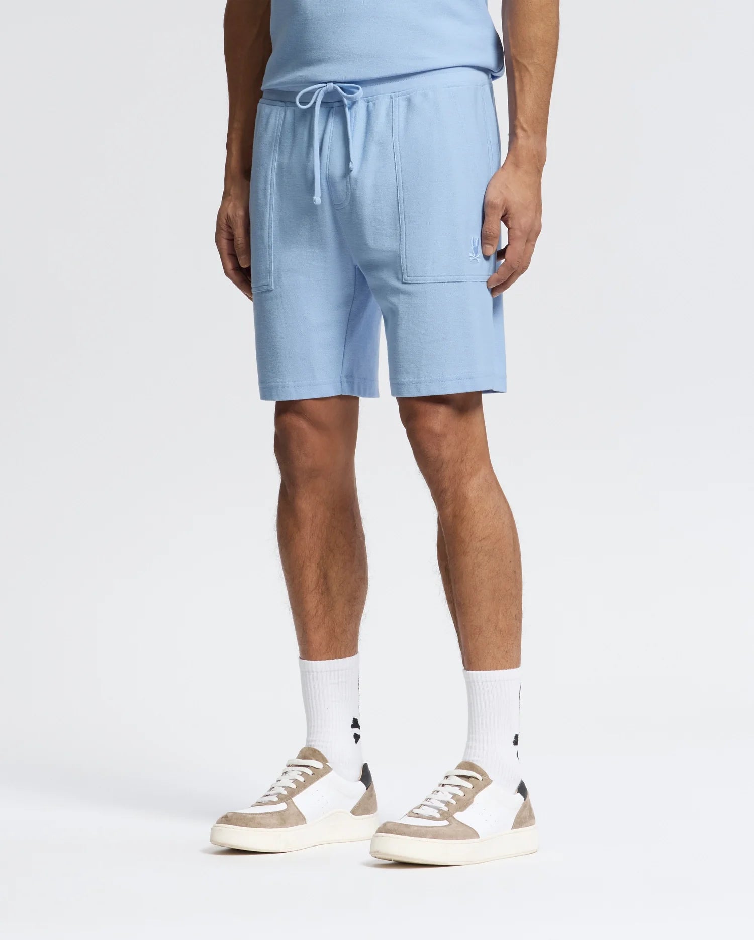Wearing Psycho Bunny's MENS TAYLOR LIGHT PIQUE SWEATSHORTS and a matching blue shirt, paired with white socks featuring a black logo and white sneakers with beige and gray highlights, creates an ideal casual-chic look against the plain white backdrop.