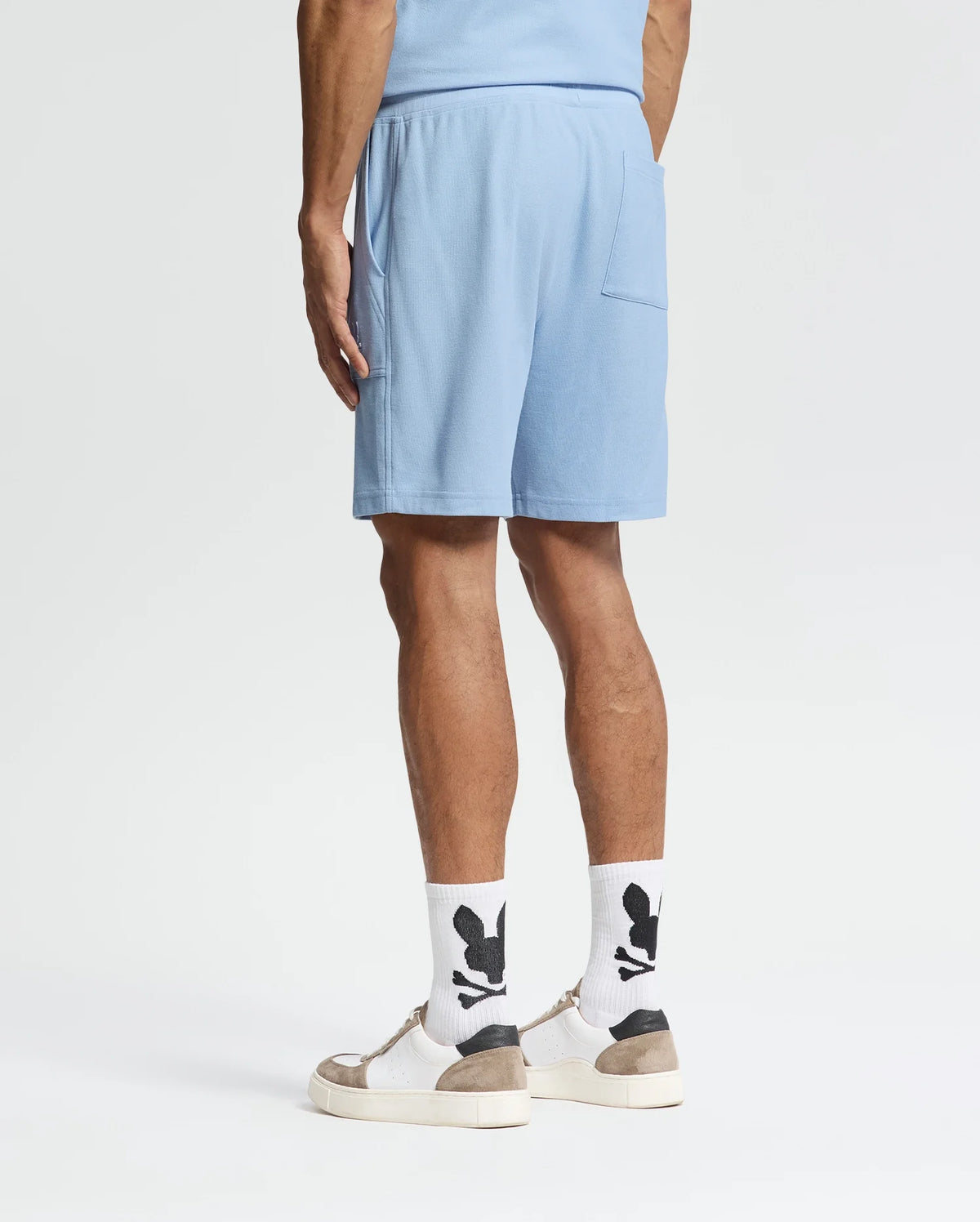 A person faces away, wearing Psycho Bunny's MENS TAYLOR LIGHT PIQUE SWEATSHORT (B6R344F200) in light blue and a coordinating top. They have white socks with a black pattern and white sneakers with beige soles against a plain white backdrop.