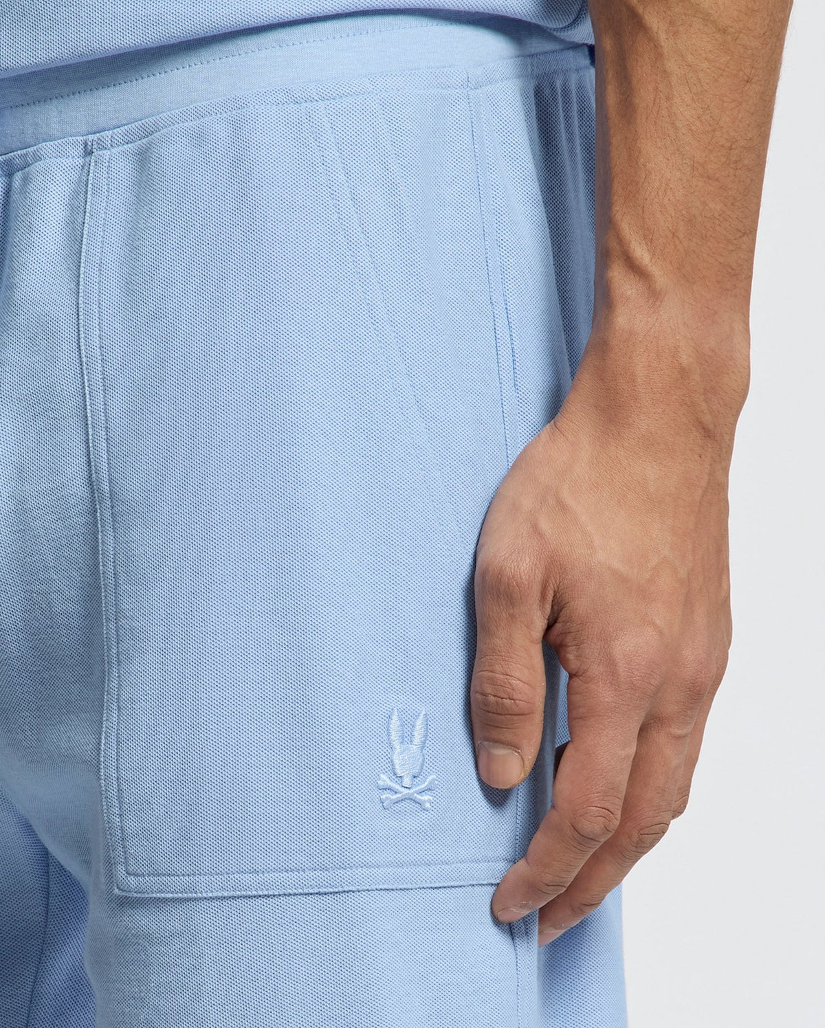 A person wears the Psycho Bunny MENS TAYLOR LIGHT PIQUE SWEATSHORT in light blue, featuring an embroidered white skull and crossbones logo on the pocket. Their right hand is near the pocket, emphasizing this casual-chic wardrobe essential's fabric and detailing.