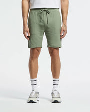 A person sports Psycho Bunny's MENS TAYLOR LIGHT PIQUE SWEATSHORT - B6R344F200 in sage green, featuring large front pockets and a drawstring waistband. They pair it with a white shirt, text-adorned white socks, and gray sneakers against a plain white background.