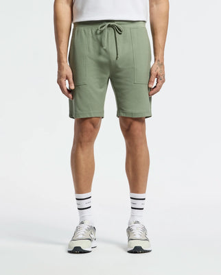 A person sports Psycho Bunny's MENS TAYLOR LIGHT PIQUE SWEATSHORT - B6R344F200 in sage green, featuring large front pockets and a drawstring waistband. They pair it with a white shirt, text-adorned white socks, and gray sneakers against a plain white background.