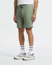 A person casually stands in lightweight sage green Psycho Bunny MENS TAYLOR LIGHT PIQUE SWEATSHORTS (B6R344F200) and a white Pima cotton shirt, completed with white socks and white-gray New Balance sneakers. Their left hand rests near the shorts pocket against a plain white background.