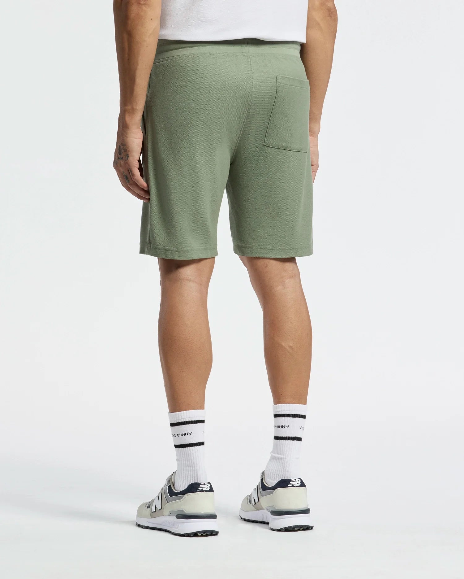Someone is standing back to the camera wearing Psycho Bunny's MENS TAYLOR LIGHT PIQUE SWEATSHORT in olive green, paired with white socks and gray sneakers. The lightweight Pima cotton shorts include a single back pocket.