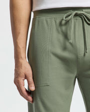 Close-up of someone wearing Psycho Bunny's Mens Taylor Light Pique Sweatshort in sage green. The drawstring shorts, made from lightweight Pima cotton, feature textured fabric and visible seams that enhance the white top against a plain backdrop.