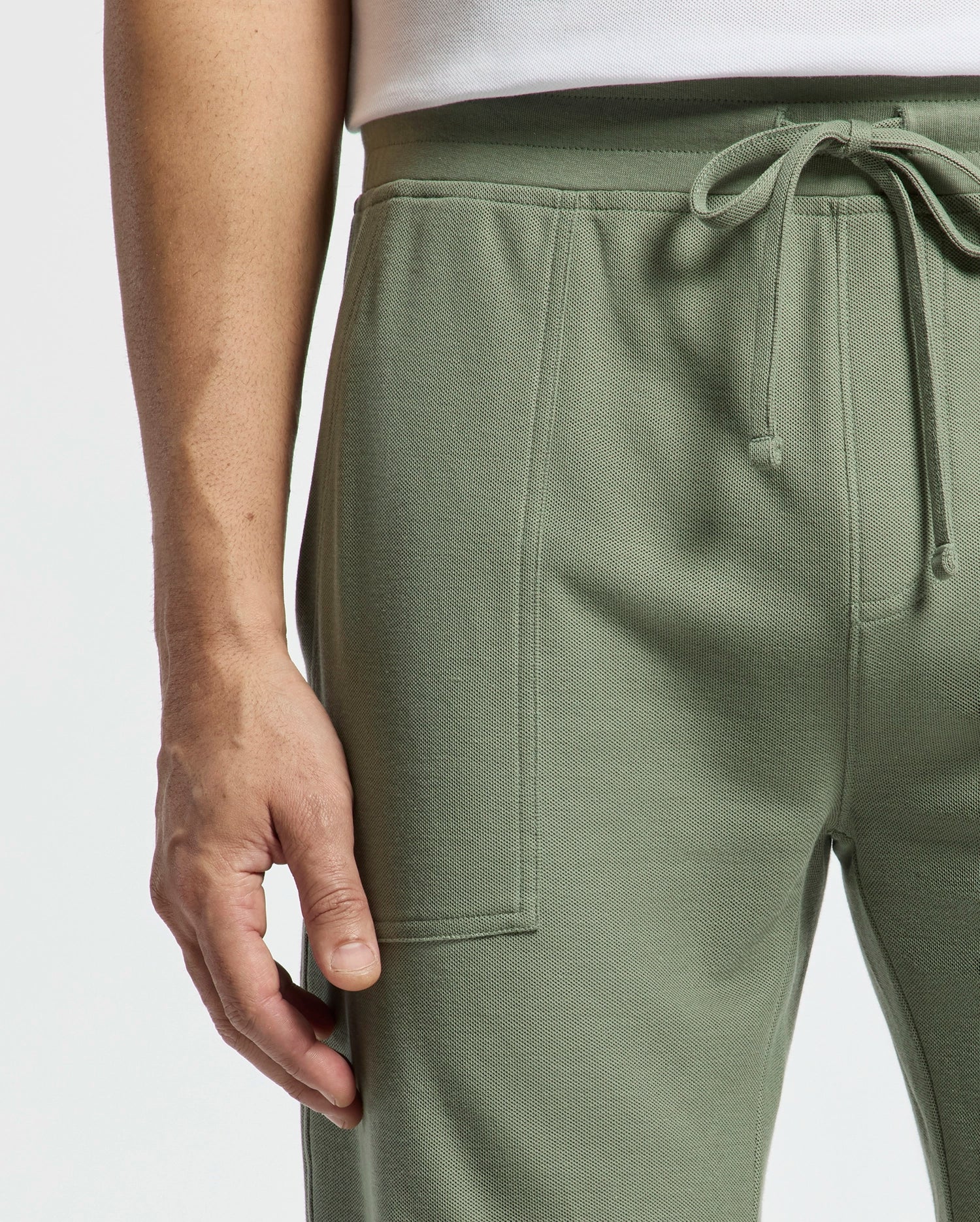 Close-up of someone wearing Psycho Bunny's Mens Taylor Light Pique Sweatshort in sage green. The drawstring shorts, made from lightweight Pima cotton, feature textured fabric and visible seams that enhance the white top against a plain backdrop.