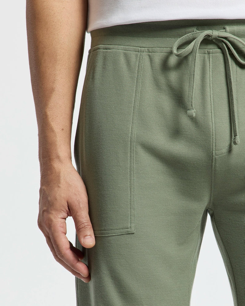 Close-up of someone wearing Psycho Bunny's Mens Taylor Light Pique Sweatshort in sage green. The drawstring shorts, made from lightweight Pima cotton, feature textured fabric and visible seams that enhance the white top against a plain backdrop.