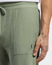A person with tattooed hands rests their hand on the green textured Psycho Bunny MENS TAYLOR LIGHT PIQUE SWEATSHORT, featuring a small embroidered logo near the pocket, while wearing a lightweight white top.