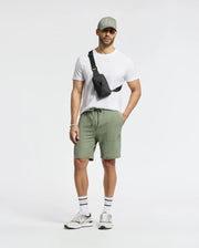 A man stands against a plain background wearing Psycho Bunny's light green MENS TAYLOR LIGHT PIQUE SWEATSHORT, made of lightweight Pima cotton. He pairs them with a white T-shirt, black crossbody bag, grey sneakers, striped socks, a watch, bracelets, and sunglasses.