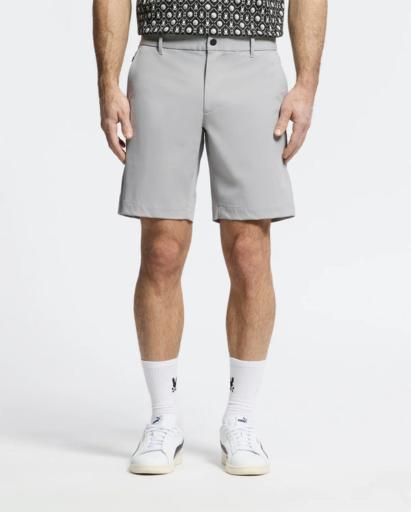 A person is wearing Psycho Bunny's MENS FRANKLIN GOLF SHORT - B6R466F200 in light gray premium nylon blend, paired with a black and white patterned shirt, white socks featuring black logos, and sleek white sneakers. The breathable athletic shorts stand out against the plain white background.