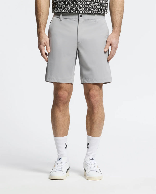 A person is wearing Psycho Bunny's MENS FRANKLIN GOLF SHORT - B6R466F200 in light gray premium nylon blend, paired with a black and white patterned shirt, white socks featuring black logos, and sleek white sneakers. The breathable athletic shorts stand out against the plain white background.