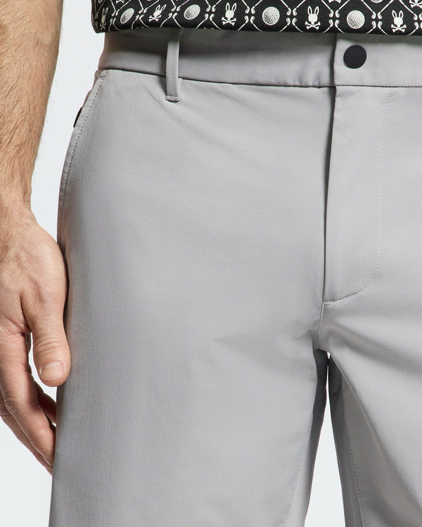 A person is shown from the waist down wearing light gray Psycho Bunny MENS FRANKLIN GOLF SHORT - B6R466F200 and a black and white patterned shirt. Their left hand is near their side, showcasing the breathable athletic material of the premium nylon blend.