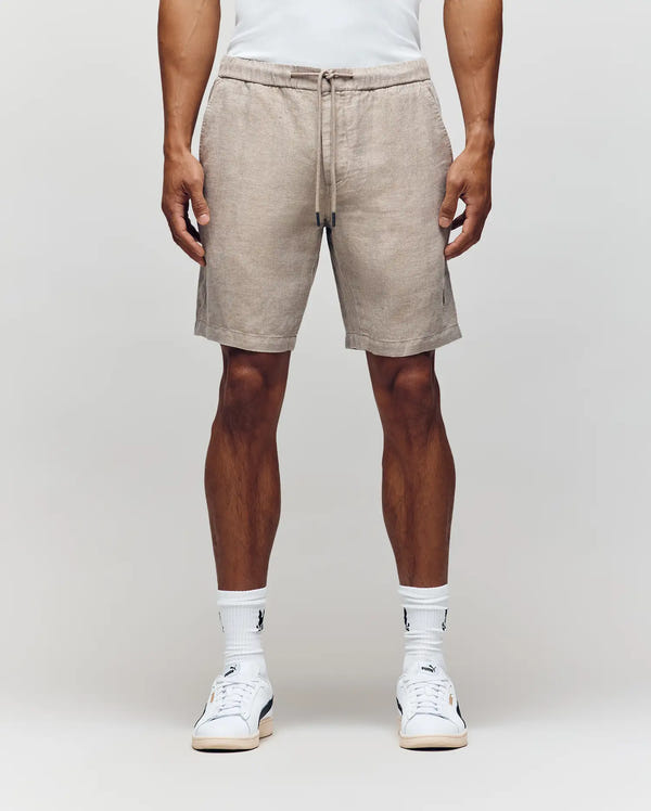 A person models Psycho Bunny’s MENS WINDCREST LINEN BLEND DRAWSTRING SHORT (B6R474C200) in light brown, paired with a white shirt, white socks, and sneakers against a plain background—an ideal look for summer style inspiration.