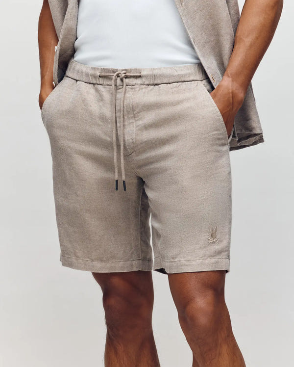A person wears Psycho Bunny's MENS WINDCREST LINEN BLEND DRAWSTRING SHORT (B6R474C200) in light beige, featuring a drawstring waist and subtle logo near the hem. Styled with a matching open shirt and white top for an easy summer look.