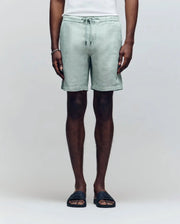 A person wears Psycho Bunny's MENS WINDCREST LINEN BLEND DRAWSTRING SHORT in light green, paired with a white top, bracelet, and black slide sandals, standing against a plain background. The image is cropped from the shoulders down.