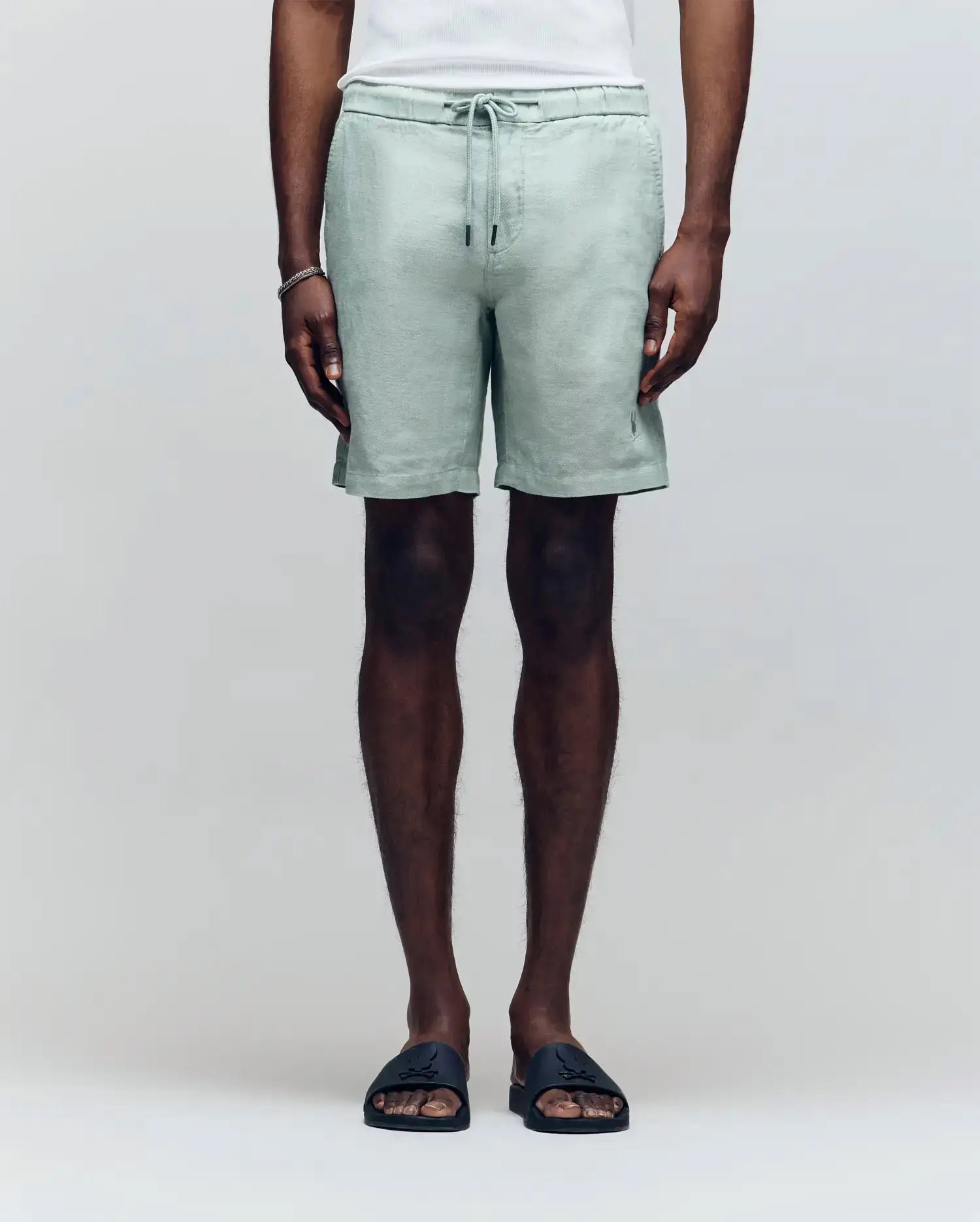 A person wears Psycho Bunny's MENS WINDCREST LINEN BLEND DRAWSTRING SHORT in light green, paired with a white top, bracelet, and black slide sandals, standing against a plain background. The image is cropped from the shoulders down.
