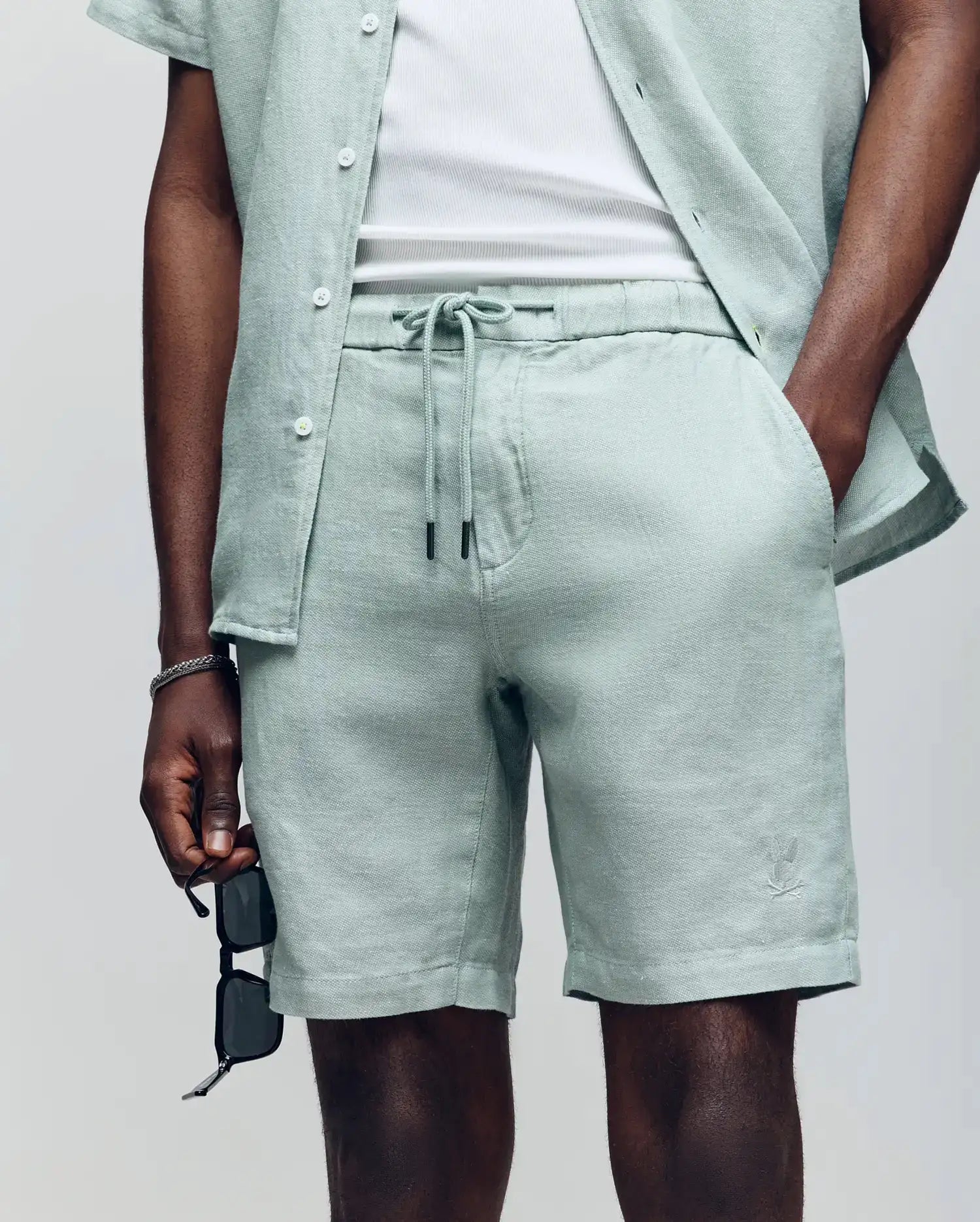 Wearing Psycho Bunny’s MENS WINDCREST LINEN BLEND DRAWSTRING SHORT (B6R474C200) and a matching light green shirt over a white T-shirt, the model poses with one hand in a pocket and black sunglasses in the other; face not shown.