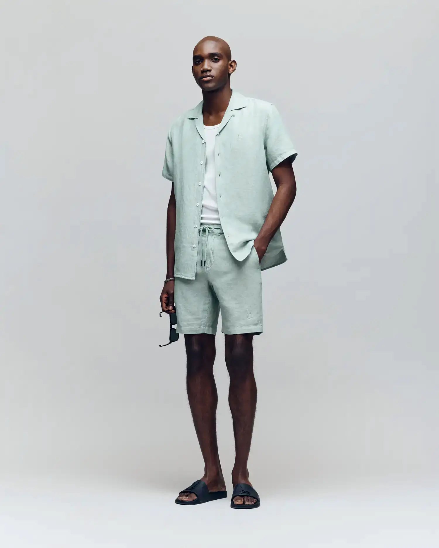 A man stands against a light gray background wearing Psycho Bunny’s MENS WINDCREST LINEN BLEND DRAWSTRING SHORT (B6R474C200), a pale green short-sleeve button-up shirt, white T-shirt, black slide sandals, and holding sunglasses.